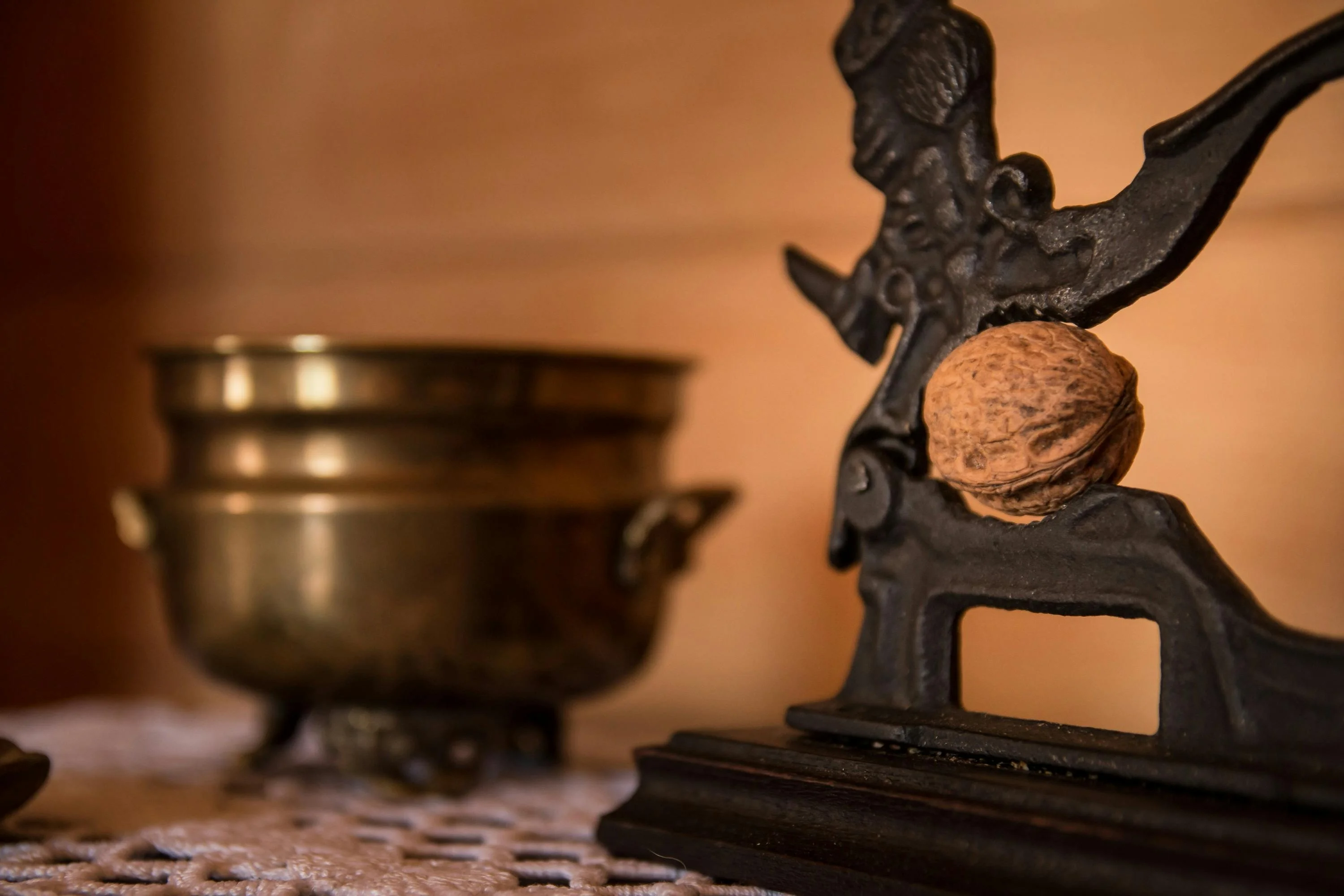 Close-up of a vintage metal nutcracker holding a walnut, with a brass bowl in the background on a lace-covered surface